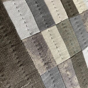 May include: A textile piece featuring a patchwork design of rectangular fabric segments in shades of dark brown, gray, and off-white. The segments are joined by visible vertical rows of dark stitching, creating a textured, linear pattern across the surface.