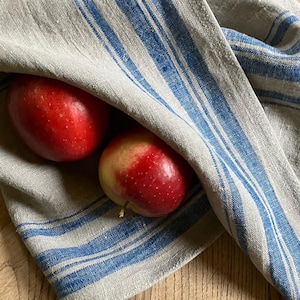 May include: A beige kitchen towel featuring vertical blue stripes of varying widths, draped over two round red apples. The fabric has a visible woven texture, and the towel is folded to partially cover the fruit while revealing the red skin.