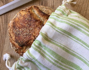 Puede incluir: Una hogaza de pan recién horneada en una bolsa de tela a rayas blancas y verdes. El pan tiene una corteza dorada y está ligeramente agrietada en la parte superior.