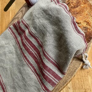 May include: A beige linen bread bag with red vertical stripes and a drawstring closure, containing a round loaf of crusty bread. Next to the bag is a glass jar of raspberry jam with a metal lid and a silver butter knife resting on top.