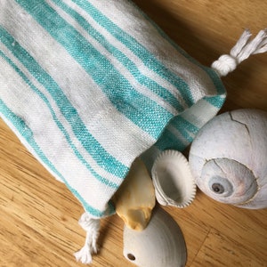 Könnte beinhalten: Eine weiße Stofftasche mit türkisfarbenen Streifen und einem Kordelzug, gefüllt mit Muscheln. Die Tasche liegt auf einer Holzoberfläche.