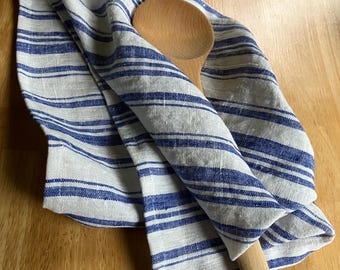 Puede incluir: Un paño de cocina de color claro con un patrón de rayas azules finas y gruesas, doblado sobre una cuchara de madera clara y lisa con un orificio circular en el extremo del mango, mostrando un diseño sencillo y funcional.