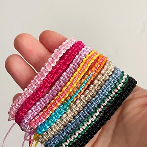 May include: A collection of colorful macrame bracelets in various shades, including pink, red, orange, yellow, green, blue, and black. Each bracelet is hand-knotted with a braided design and adjustable string closure. The bracelets are displayed on a hand.