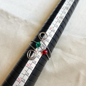 May include: A handmade wire ring with a swirl design, adorned with green, clear, and red beads. The ring is shown on a black ring sizer, with measurements visible in inches. The ring's design is simple yet elegant.