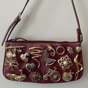 May include: Burgundy leather shoulder bag adorned with an assortment of gold-toned brooches. The bag features a long strap and a zipper closure. Brooches include floral, heart, and animal designs.