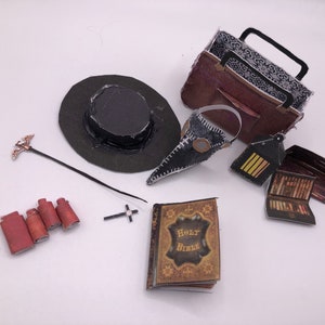 Op de afbeelding: Een verzameling miniatuurvoorwerpen, waaronder een zwarte hoge hoed, een pestmeestermasker, een bruine tas, een staf met een vogel, vier kleine rode rollen, een kruis, een lantaarn, een klein boek en een miniatuur Bijbel met gouden letters.