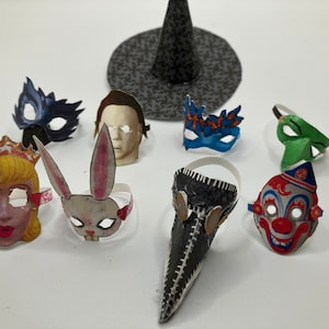 Op de afbeelding: Een verzameling miniatuur papieren maskers waaronder een prinses, een konijn, een pestmeester, een clown, een menselijk gezicht en diverse maskerade stijlen, vergezeld door een kleine zwarte heksenhoed met een patroon op een effen ondergrond.