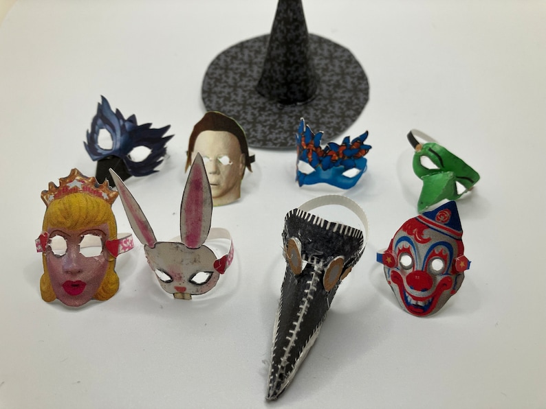 Op de afbeelding: Een verzameling miniatuur papieren maskers waaronder een prinses, een konijn, een pestmeester, een clown, een menselijk gezicht en diverse maskerade stijlen, vergezeld door een kleine zwarte heksenhoed met een patroon op een effen ondergrond.