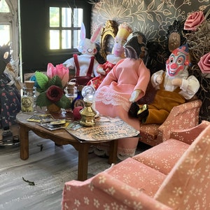 Op de afbeelding: Een miniatuur poppenhuis woonkamer met een houten salontafel met een spiritbord, kleine glazen flesjes en een koperen lamp. Verschillende poppen zitten op een bank en stoelen, waaronder figuren met maskers, een clown en een konijn.
