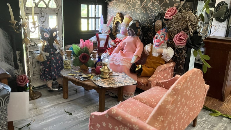 Op de afbeelding: Een miniatuur poppenhuis woonkamer met een houten salontafel met een spiritbord, kleine glazen flesjes en een koperen lamp. Verschillende poppen zitten op een bank en stoelen, waaronder figuren met maskers, een clown en een konijn.