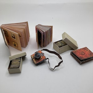 May include: A collection of miniature items including two small books with internal pages, a tiny orange and black camera with a strap, two rectangular boxes filled with small paper cards, and a square box with a red floral patterned lid.