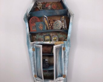 May include: A coffin-shaped paper craft display shelf with a light blue distressed finish. The interior features multiple shelves decorated with floral wallpaper and various miniature paper cutouts including a skull, hand, and camera. Little Paper Worlds.