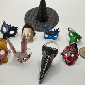 Op de afbeelding: Een verzameling miniatuur papieren maskers waaronder een prinses, een konijn, een pestdokter, een clown en diverse karaktergezichten, vergezeld door een kleine zwart gepatroonde heksenhoed op een effen witte ondergrond.