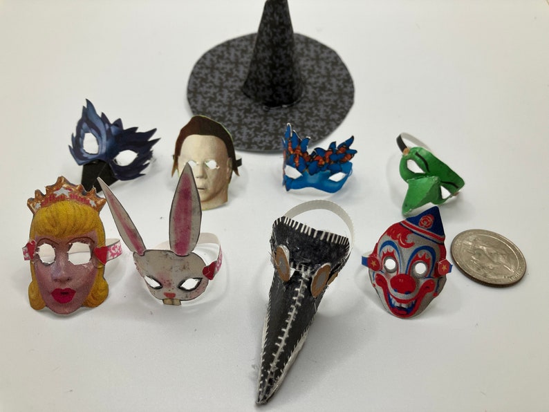 Op de afbeelding: Een verzameling miniatuur papieren maskers waaronder een prinses, een konijn, een pestdokter, een clown en diverse karaktergezichten, vergezeld door een kleine zwart gepatroonde heksenhoed op een effen witte ondergrond.