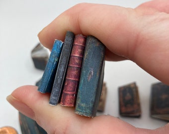 May include: A collection of four miniature books held together, featuring spines in blue, dark grey, reddish-brown with horizontal ridges, and a weathered dark teal. These tiny volumes are displayed alongside other small book items and a copper penny.