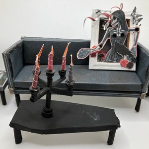 May include: A miniature furniture set featuring a dark gray sofa, a black coffin-shaped coffee table with a black candelabra holding five thin candles, a small side table, and a white framed wall art piece depicting a skeletal figure with a raven.