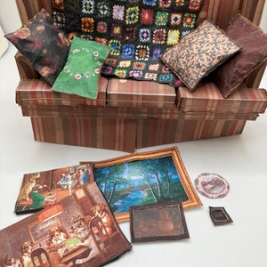 Può includere: Un divano in miniatura di cartone con rivestimento a righe marroni, dotato di una coperta nera a motivi quadrati e quattro piccoli cuscini. Sotto si trovano diverse stampe artistiche in miniatura, tra cui scene di cani che giocano a carte.