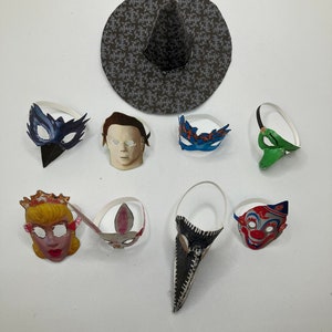 Op de afbeelding: Een verzameling papieren maskers en een heksenhoed. De set bevat een donkerblauw vogelmasker, een bleek menselijk gezichtsmasker, een blauw met oranje masker, een groen masker, een blond karaktermasker, een konijnenmasker, een lang zwart snavelmasker en een clownmasker.