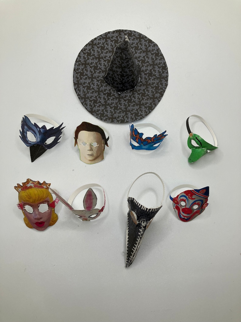 Op de afbeelding: Een verzameling papieren maskers en een heksenhoed. De set bevat een donkerblauw vogelmasker, een bleek menselijk gezichtsmasker, een blauw met oranje masker, een groen masker, een blond karaktermasker, een konijnenmasker, een lang zwart snavelmasker en een clownmasker.