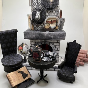Op de afbeelding: Een miniatuur poppenhuis-scène met een open haard met een ramschedel, een ronde tafel met een theepot en theekopjes, twee zwarte gecapitonneerde stoelen, een wereldbol met bloemmotieven en een open boek met een silhouet-illustratie.