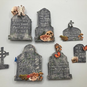 Op de afbeelding: Een verzameling van zes grijze grafsteen-vormige decoraties met diverse gedrukte grafschriften, schedels, vlinders, kikkers en paddenstoelen. De grafstenen tonen teksten zoals Here lies John Yeast, Rest in Peace cousin Huet, I told you I was sick en Here lies Lester Moore.