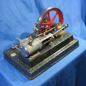 Op de afbeelding: Een kleine, zwarte en zilveren model stoommachine met een rood wiel. De machine staat op een zwarte basis en bevindt zich op een blauw oppervlak.