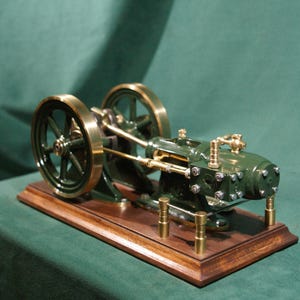 Op de afbeelding: Een gedetailleerd model van een stoommachine, voornamelijk donkergroen met messing accenten. De wielen en andere componenten zijn messingkleurig. Het is gemonteerd op een houten voet. Het model is een gedetailleerde weergave van een industriële machine.