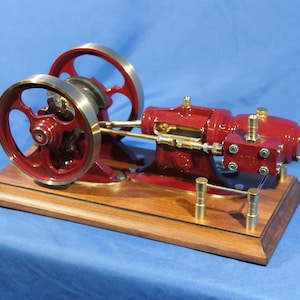 Peut inclure: Une machine à vapeur miniature rouge et laiton sur une base en bois. Le moteur a deux volants et un piston visible. Le moteur est monté sur une base en bois avec des accents en laiton.