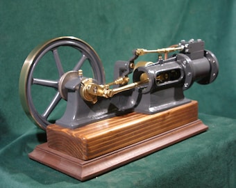 May include: A small, gray cast iron steam engine with brass accents, mounted on a wooden base. The engine has a flywheel and a connecting rod.