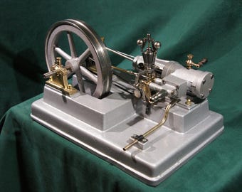 May include: A small, silver-coloured model steam engine with a brass flywheel and connecting rods. The engine is mounted on a silver base and has a black pipe running to the side.