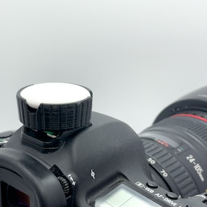 May include: A black DSLR camera with a white diffuser attached to the hot shoe. The camera lens is a 24-105mm zoom lens.