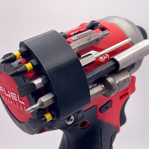 May include: A red and black cordless drill with a black 3D printed attachment that holds multiple drill bits and screwdriver bits. The drill has the text "FUEL BRUSHLESS" on the side.