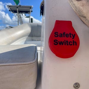 May include: A red safety switch with the words "Safety Switch" in black text. The switch is mounted on a white surface, likely on a boat. The background shows boat seating and a blue sky.