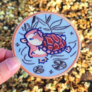 May include: Embroidered patch featuring a pink and white cartoon platypus with a red and brown turtle shell, swimming in a light blue pond with lily pads. The patch has a light brown border and is held by a hand.