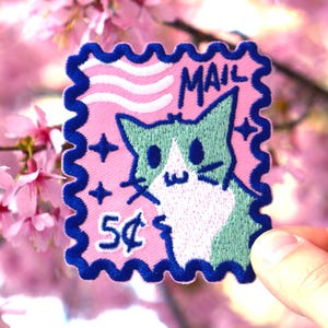 May include: A pink and blue embroidered patch in the shape of a postage stamp featuring a cartoon cat. The patch has the word "MAIL" and the number "5¢" on it. The background is a blurred image of pink cherry blossoms.
