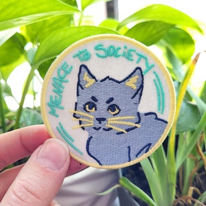 May include: A white embroidered patch with a blue cat with yellow eyes and ears. The text "Menace to Society" is written in green around the cat.