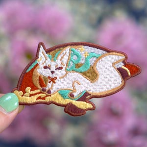 May include: Embroidered patch featuring a white fox with a red bow, resting on a bed of gold and brown. The fox has teal and gold accents, with a brown border. The patch is held against a blurred floral background.