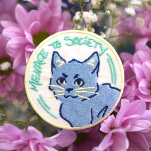 May include: Embroidered patch featuring a blue cat with yellow eyes and whiskers, surrounded by the text "MENACE TO SOCIETY" in green. The patch is set against a backdrop of pink flowers and white baby's breath.
