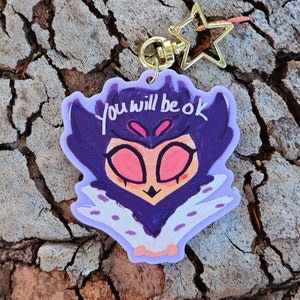 May include: Purple and white cartoon character keychain with the text "you will be ok" on a light purple background. The keychain has a gold star charm.