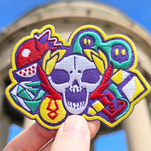 May include: A colorful embroidered patch featuring a skull with a purple background. The skull is surrounded by various shapes and designs, including a red and yellow lightning bolt, a green and white diamond, and a yellow and white ghost.