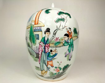 Puede incluir: Jarrón de porcelana blanca con tapa, decorado con una escena pintada a mano de una mujer y tres niños en un jardín. La escena presenta una casa, árboles, flores y un estanque de loto. Los colores son vibrantes e incluyen tonos de verde, azul, rosa y rojo.