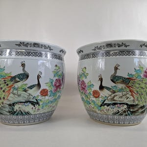 Op de afbeelding: Twee bijpassende witte keramische plantenbakken met een gedetailleerd pauw- en bloemmotief. De plantenbakken hebben een brede rand met een decoratieve rand en een kleurrijke illustratie van pauwen, bloemen en bladeren.