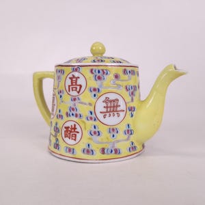 May include: A yellow ceramic teapot featuring a repeating blue and red floral pattern and three circular medallions containing red Chinese characters. The teapot has a matching lid with a small knob, a curved handle, and a tapered spout.