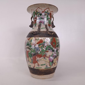 Peut inclure: Un vase en céramique avec une finition émaillée craquelée, présentant des scènes peintes à la main de personnages à cheval et des bandes brun foncé ornées. Le vase comprend deux anses sculptées en forme de créatures mythiques.