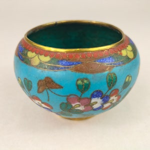 May include: A small, round, blue and white cloisonné bowl with floral designs. The bowl has a gold rim and a green interior.