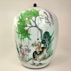 May include: A white porcelain jar with a lid, decorated with a green and brown landscape scene featuring figures and trees. The jar is painted with green, brown, pink, and black colors.