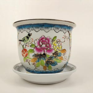 Pode incluir: Vaso de cerâmica branco com um design floral azul e dourado com um pássaro, flores e folhas. O vaso tem um pires correspondente.