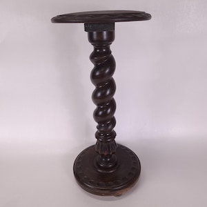 May include: A dark brown wooden pedestal stand featuring a circular top with a scalloped edge, a central column with a prominent spiral twist design, and a matching circular base with a decorative carved border detail at the bottom.