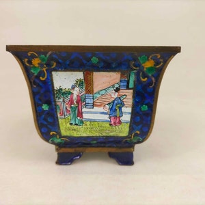 Puede incluir: Un pequeño macetero de esmalte azul y dorado con una escena cuadrada de dos figuras en un jardín. El macetero tiene tres patas y un borde festoneado.
