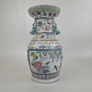 Pode incluir: Um vaso de cerâmica alto e ornamentado com boca larga e duas alças. O vaso é branco com desenhos florais e de pássaros coloridos, incluindo flores rosa, amarelas e azuis. A borda e as alças têm um detalhe verde. O vaso tem cerca de 30 cm de altura.